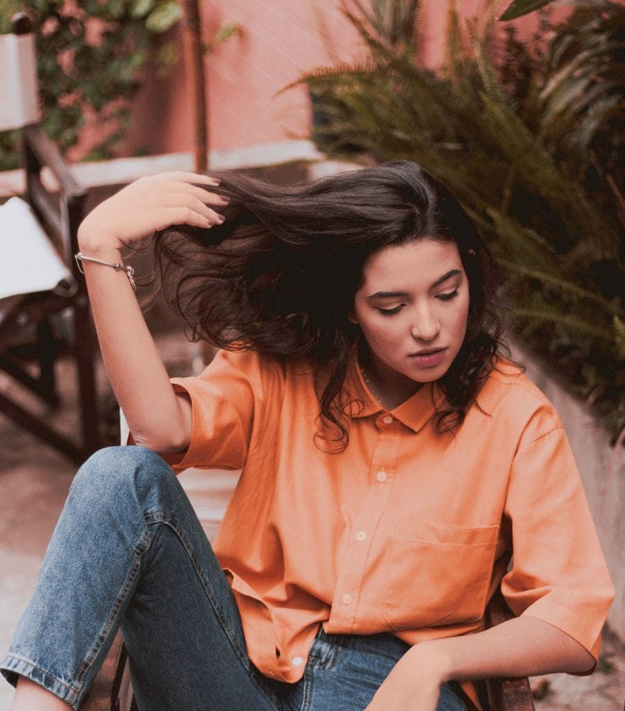 Woman in orange shirt and blue jeans running fingers through her hair